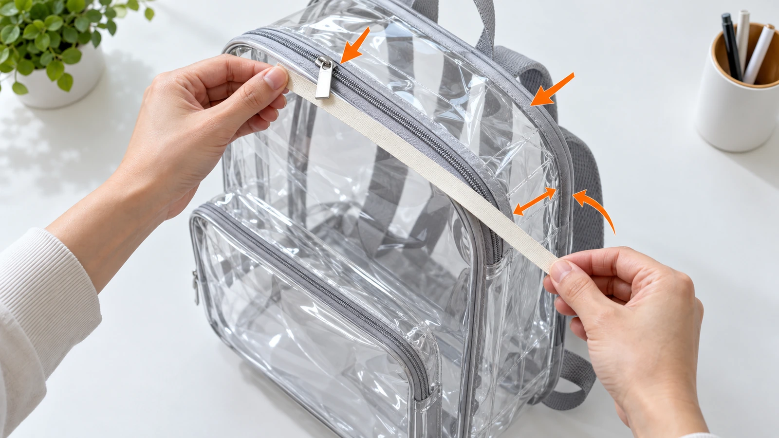 Mains mesurant, avec un simple mètre ruban, la hauteur, la largeur et la profondeur de soufflet hors tout d'un sac à dos transparent en PVC, avec des flèches indiquant que les tirettes de fermeture éclair et le ruban de finition comptent dans la taille